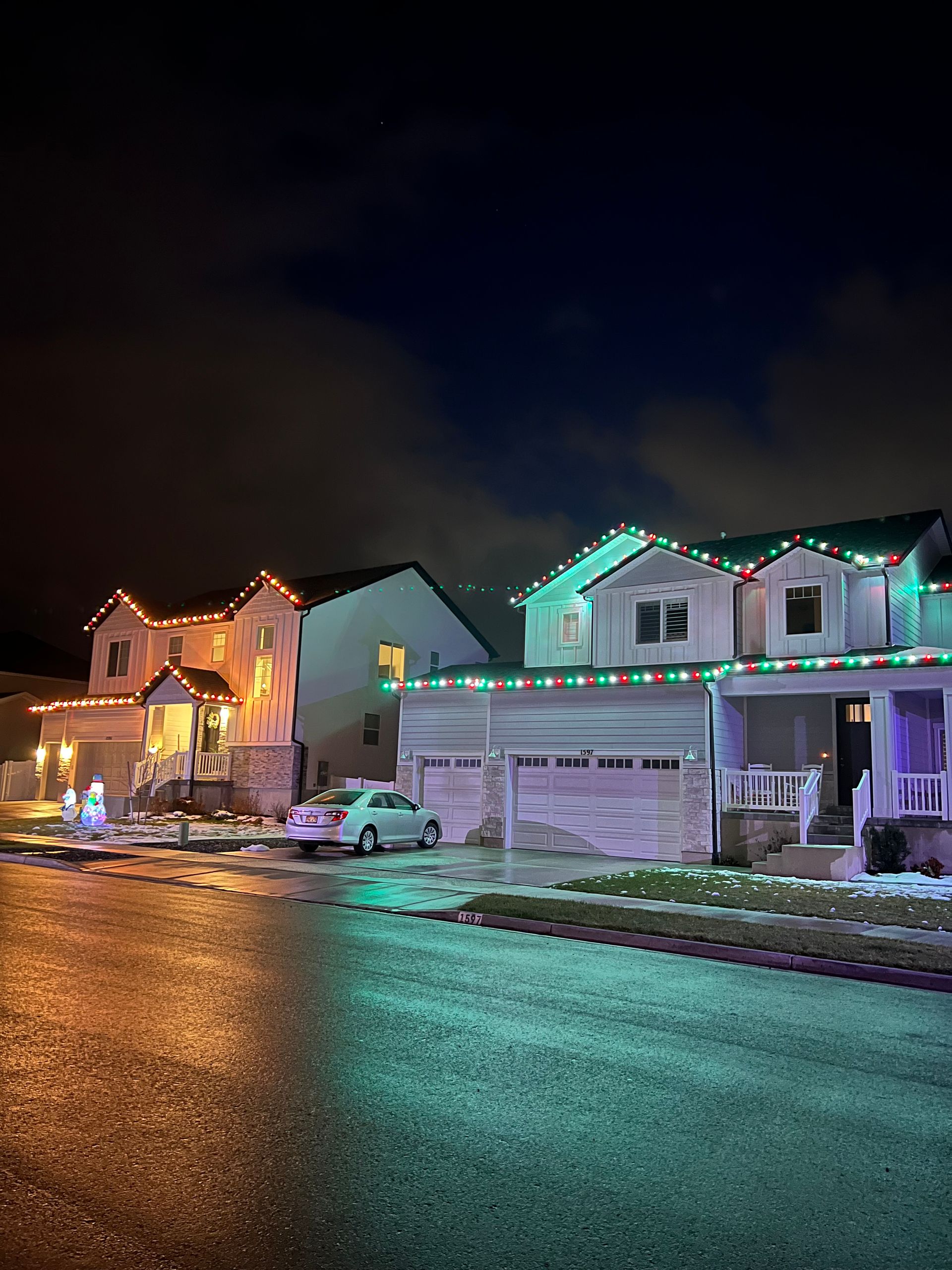 A cozy suburban house at night, adorned with white holiday lights along the roof. Warm light glows from windows, and a two-car garage is visible.