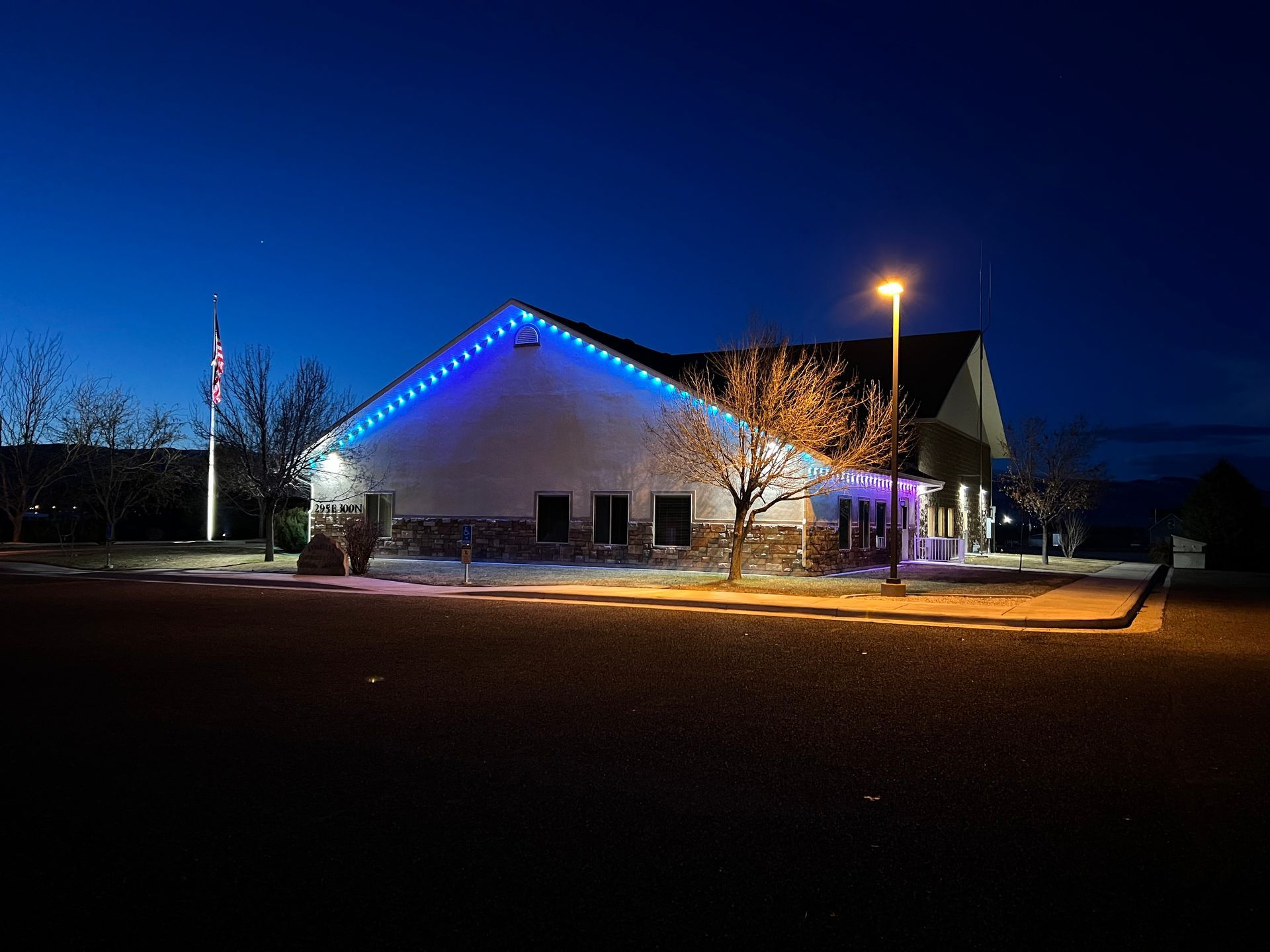 A building at dusk is adorned with blue lights along the roof, next to a flagpole. Streetlights cast a warm glow on the otherwise dark surroundings.