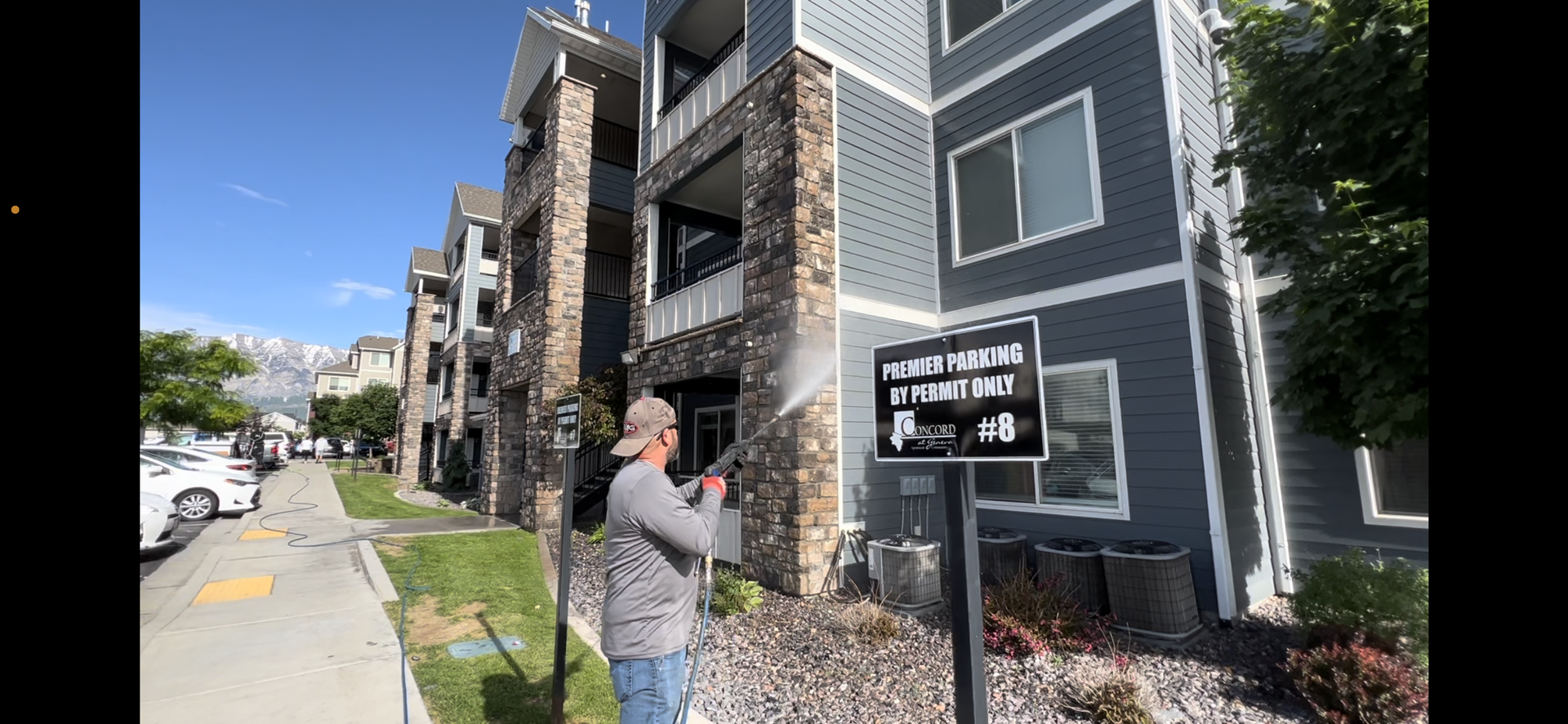 A person pressure washes in front of a modern apartment building with stone and siding. A sign reads 