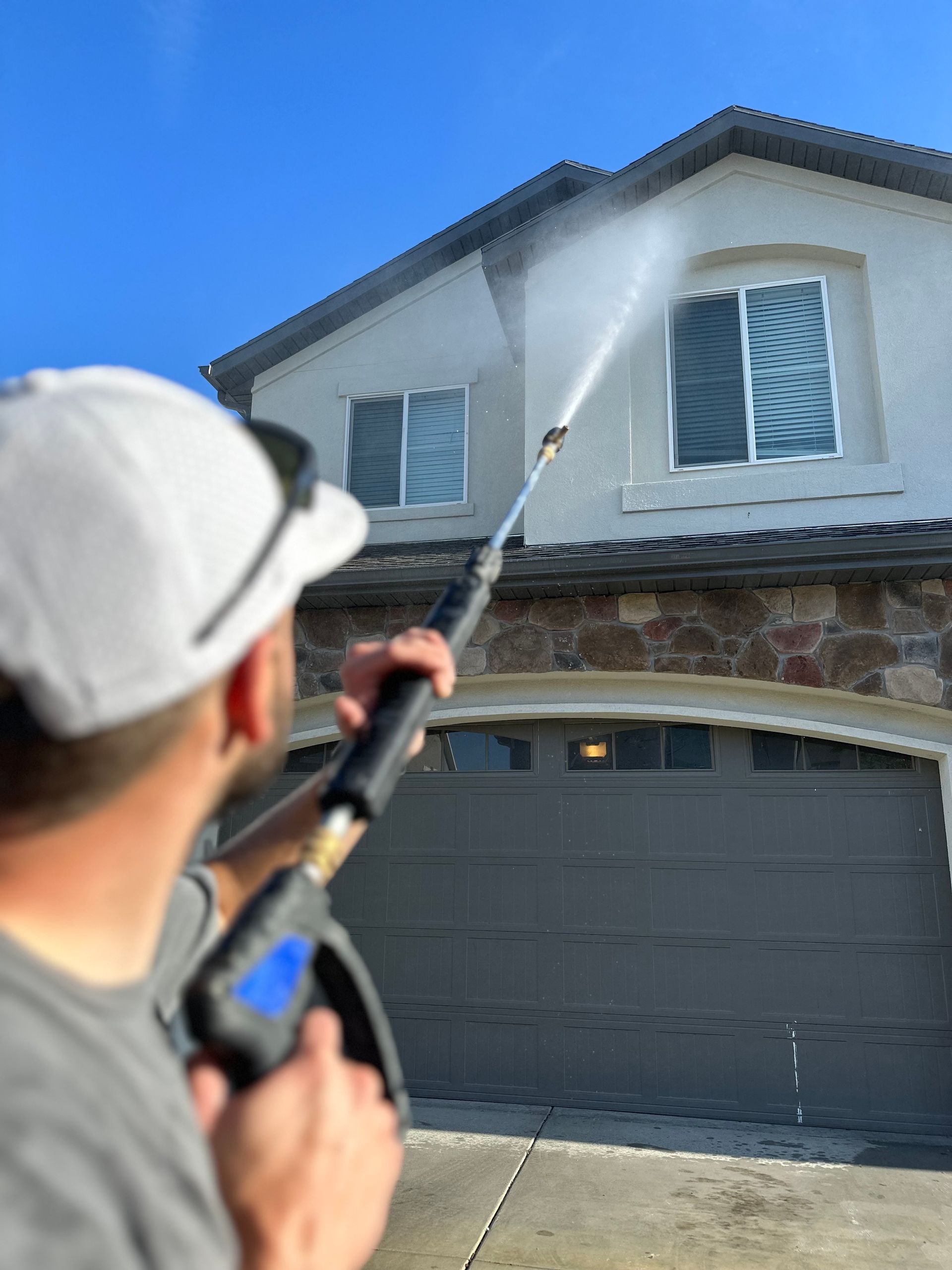 A person in a cap uses a power washer to clean a home's stone and stucco facade. The spray reaches a second-story window under a clear blue sky.