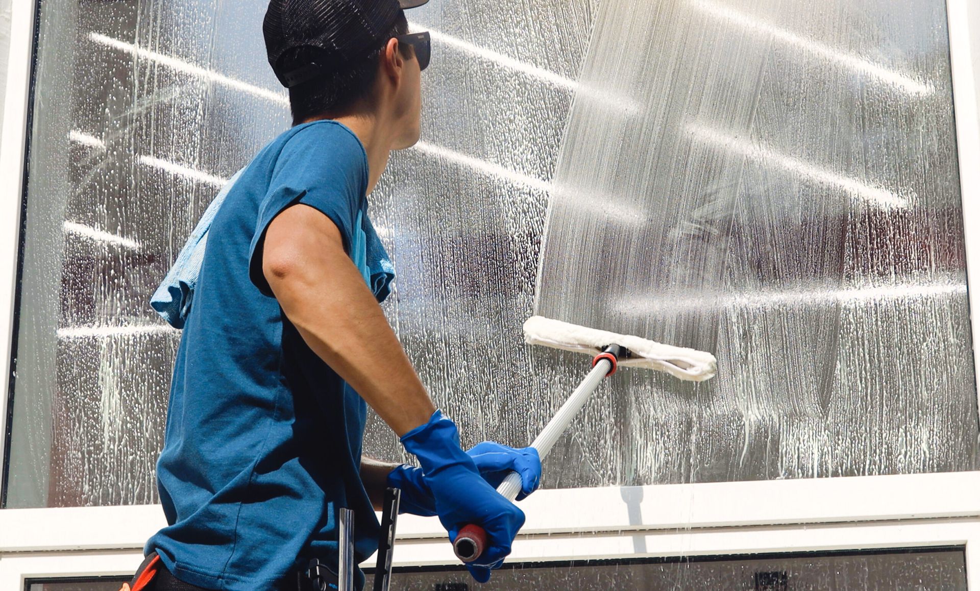 A person wearing a blue shirt and gloves cleans a large window with a squeegee, creating patterns of suds on the glass. The scene is bright and industrious.