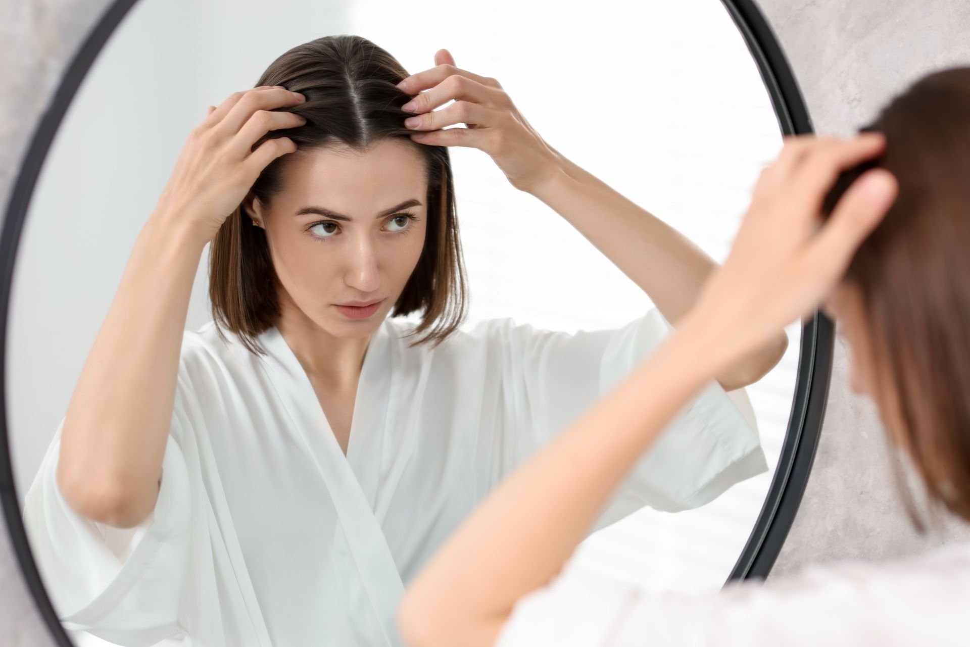 Woman examining her hair in a mirror while adjusting her part, wearing a white robe.