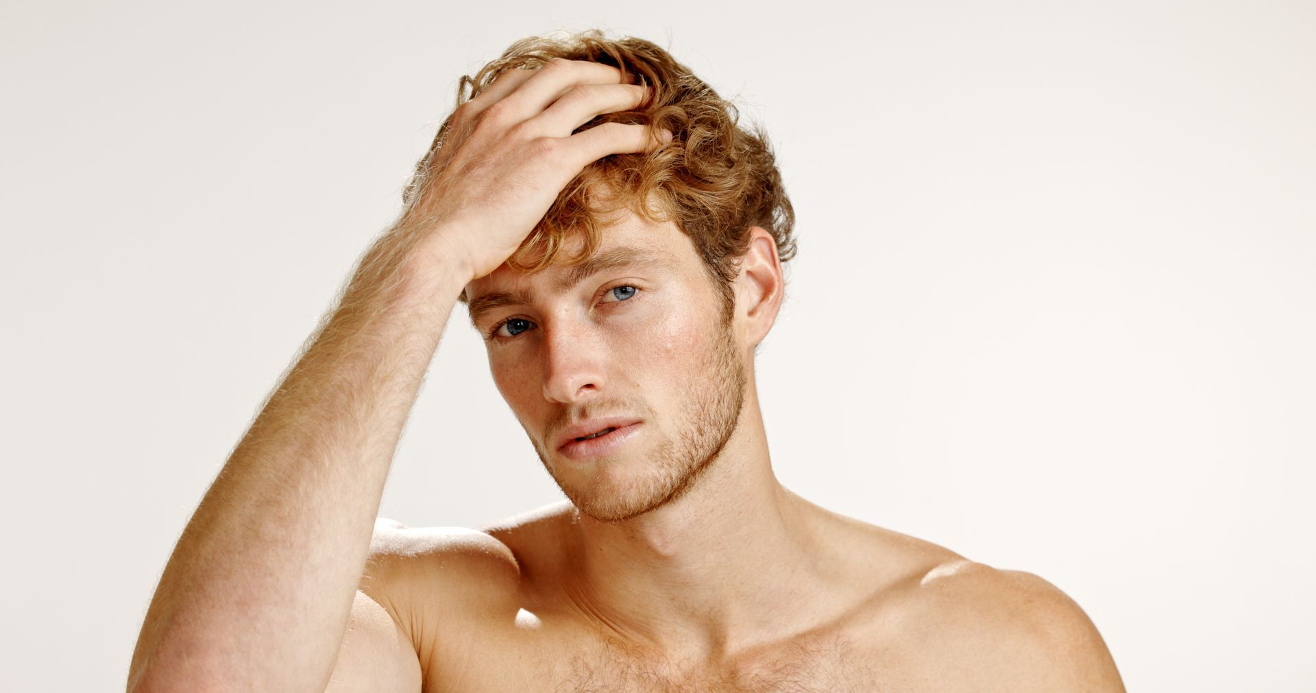 A person with light brown curly hair rests their hand on their forehead against a plain, light background.
