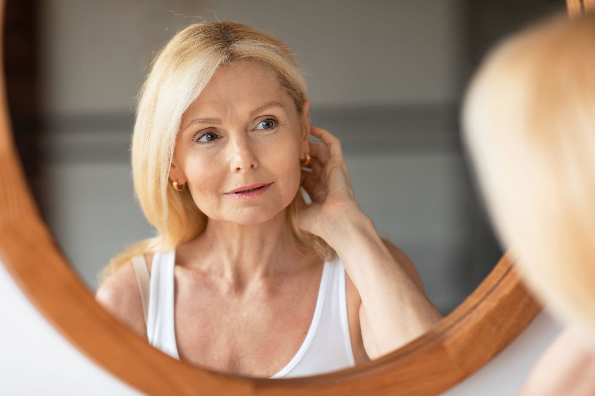 A fair-haired person looks into a round mirror, touching their hair while wearing a white tank top.