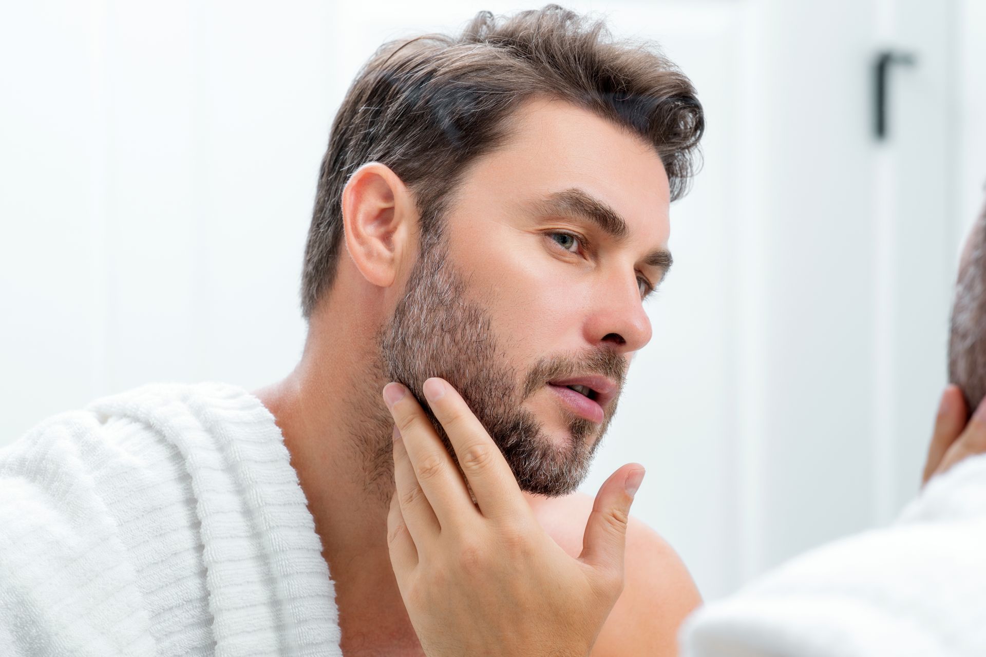 A person with short, dark facial hair wearing a white towel looks in a mirror and touches their cheek.