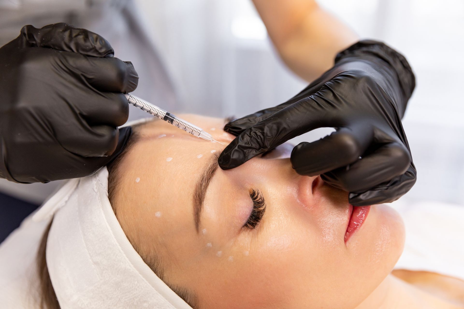 A person receiving a cosmetic facial injection from a professional wearing black gloves in a clinical setting.