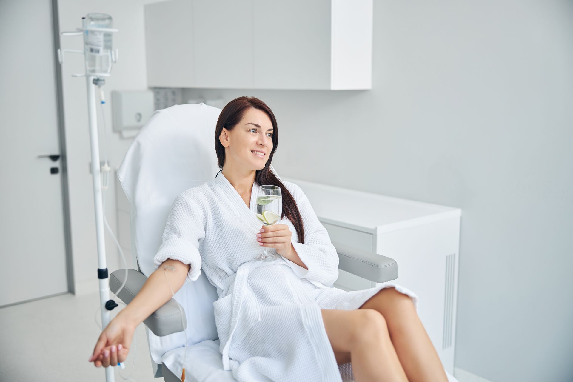Woman smiling in a hospital chair, holding a drink and resting with an IV beside her.