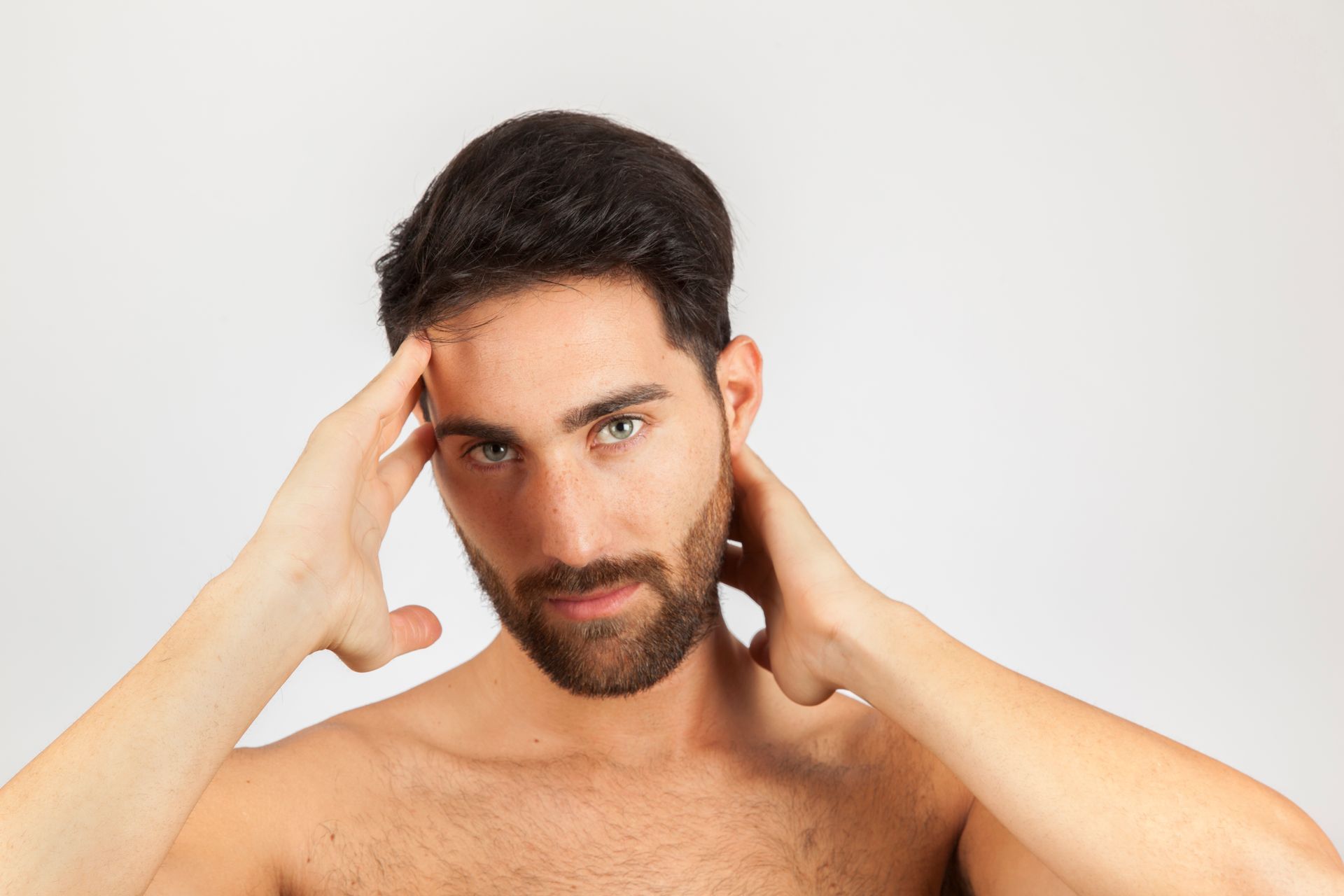 A shirtless man with a beard is holding his head with his hands.