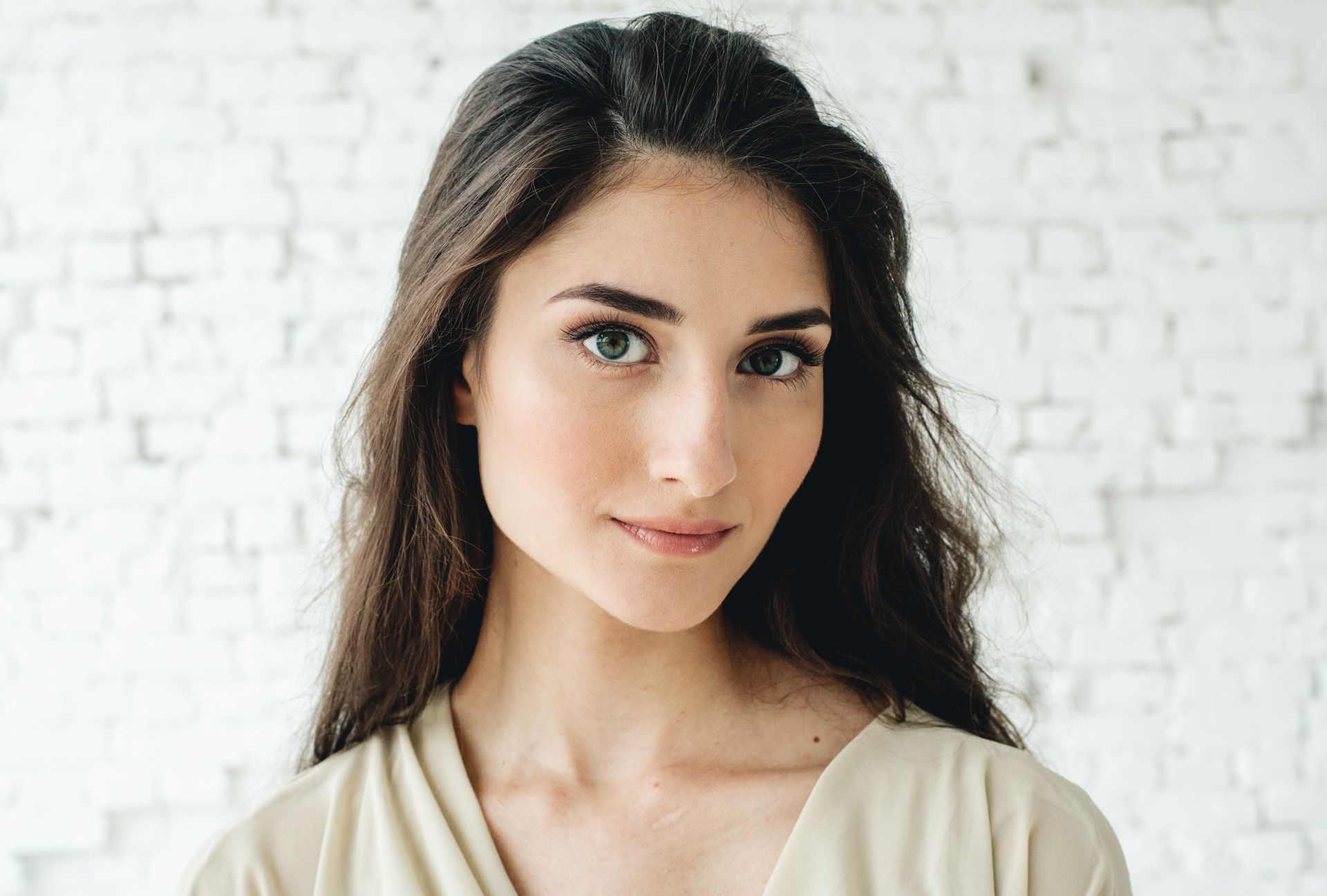 A woman with long hair is standing in front of a white brick wall and looking at the camera.