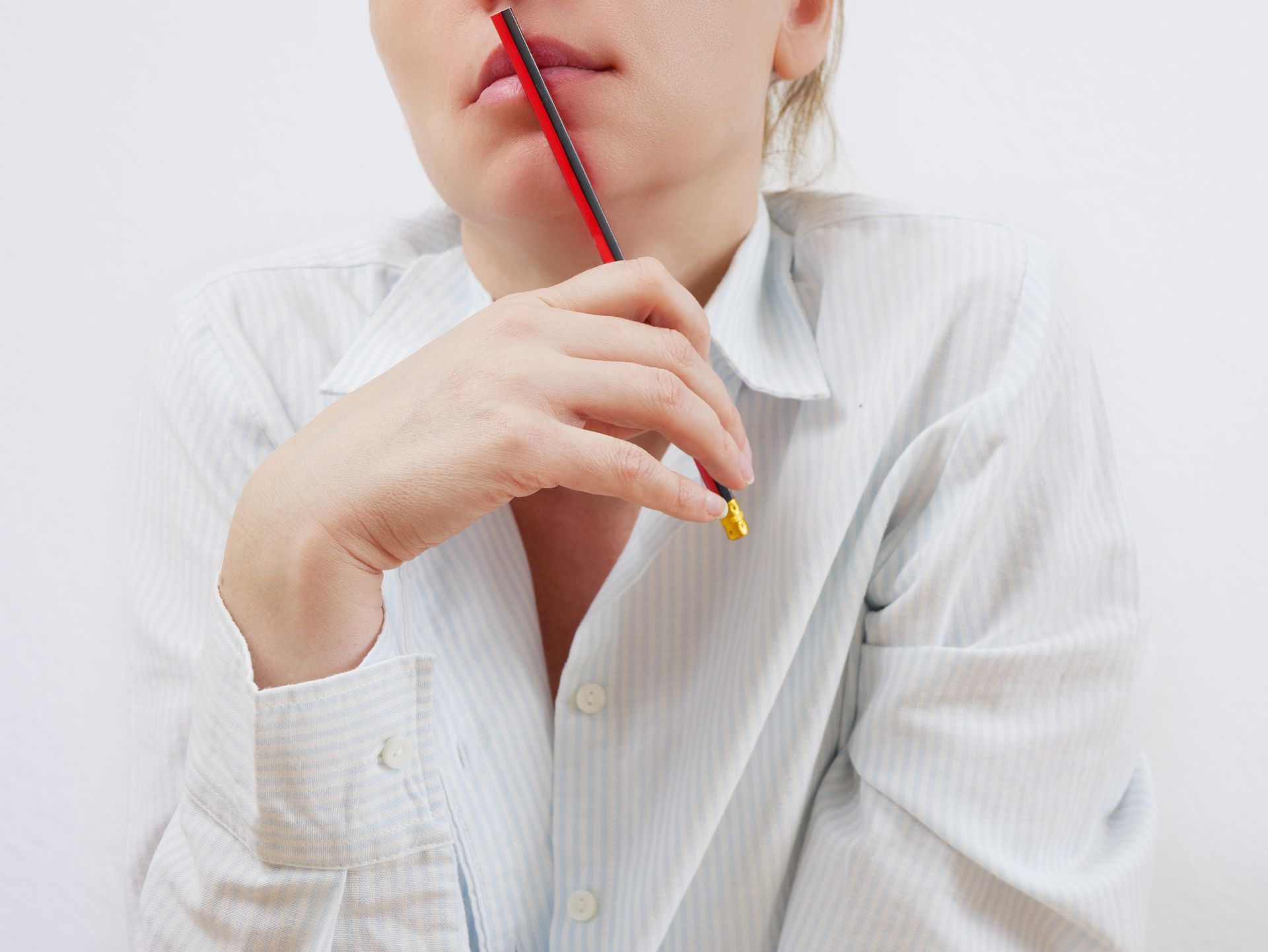 Person holding a pencil while thinking about a decision. Person in white shirt holding a red pencil near their lips, resting their chin on a hand.