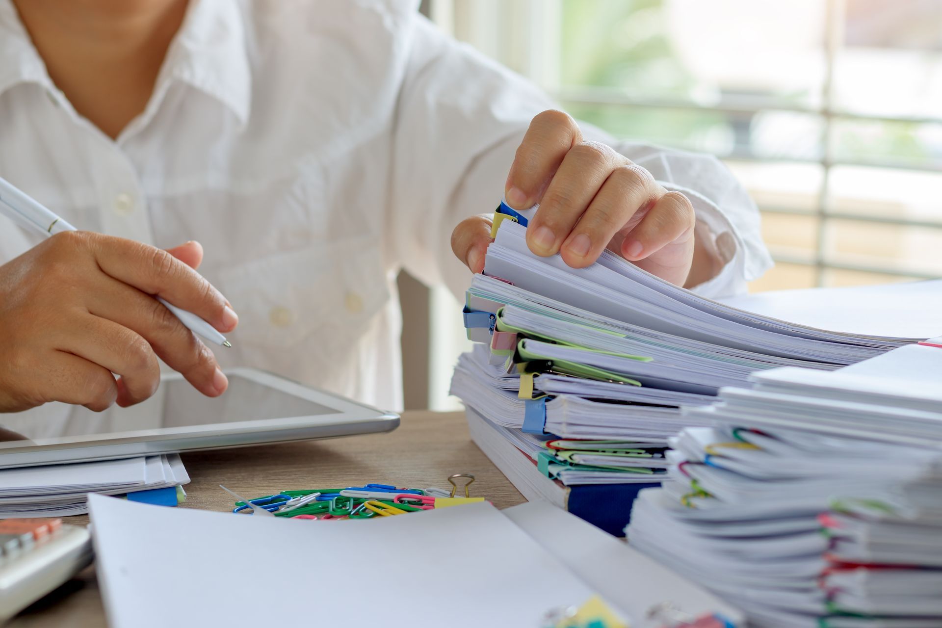 Teacher grading exam papers at desk with QME Test Prep materials. Person using a tablet beside a large stack of papers and folders on a desk