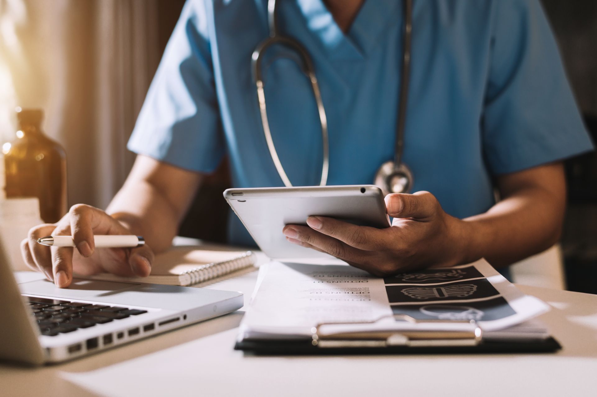 Healthcare professional using a tablet and laptop while reviewing patient records at a desk.