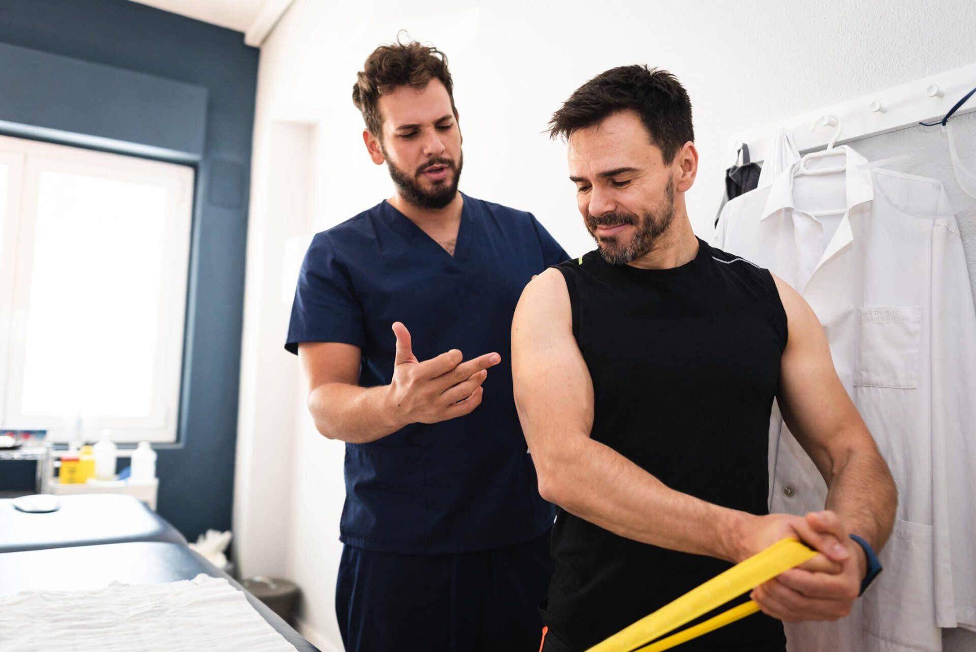 Two men in a bright room, one helping the other stretch a yellow resistance band.