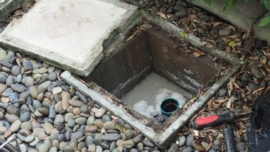 Open drainage box with rocks, a concrete cover, and a red-handled tool beside it