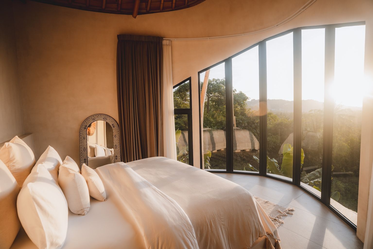 Sunlit bedroom with white bed and curved floor-to-ceiling windows overlooking a scenic view