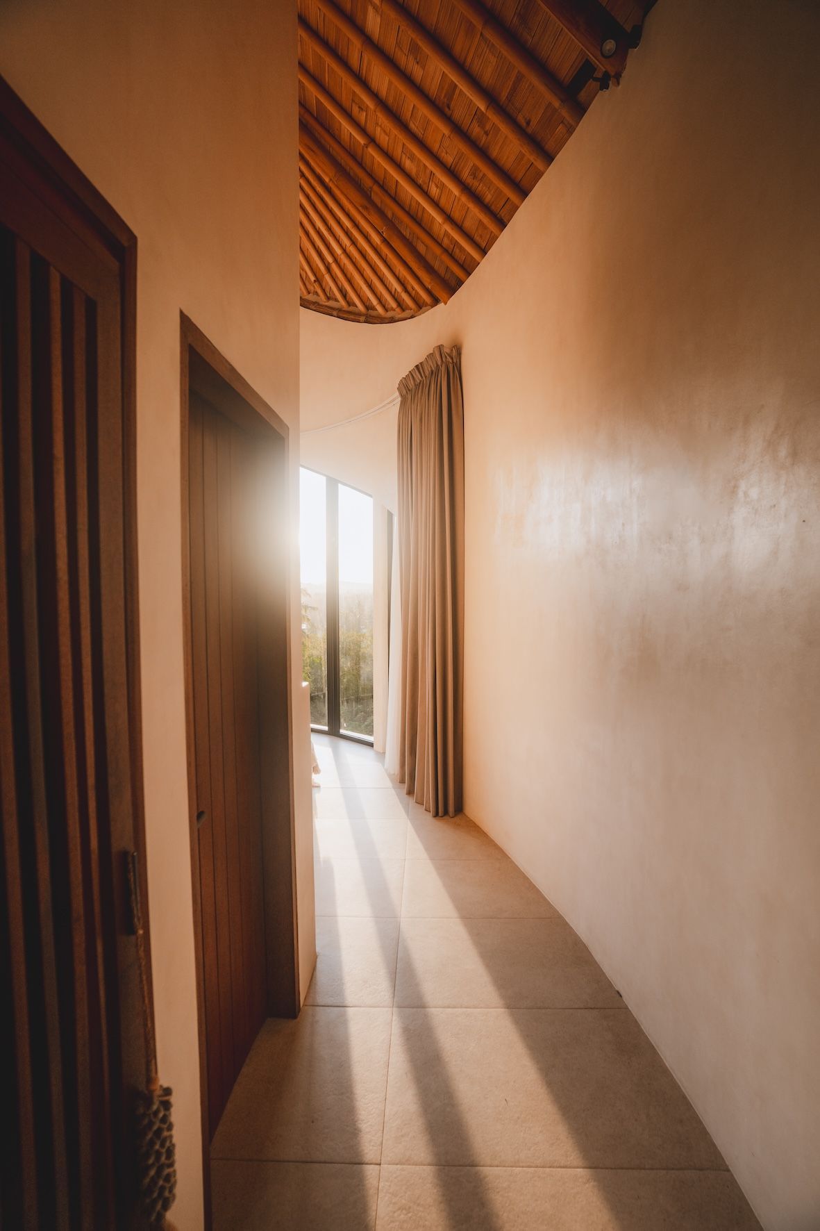 Sunlit narrow hallway with tiled floor, wooden ceiling, and open doorway at the end