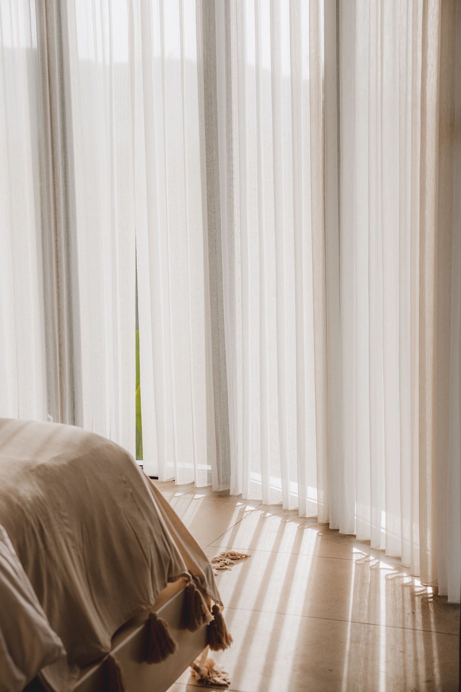 Sunlit bedroom corner with sheer white curtains, a bed, and light streaming onto the floor