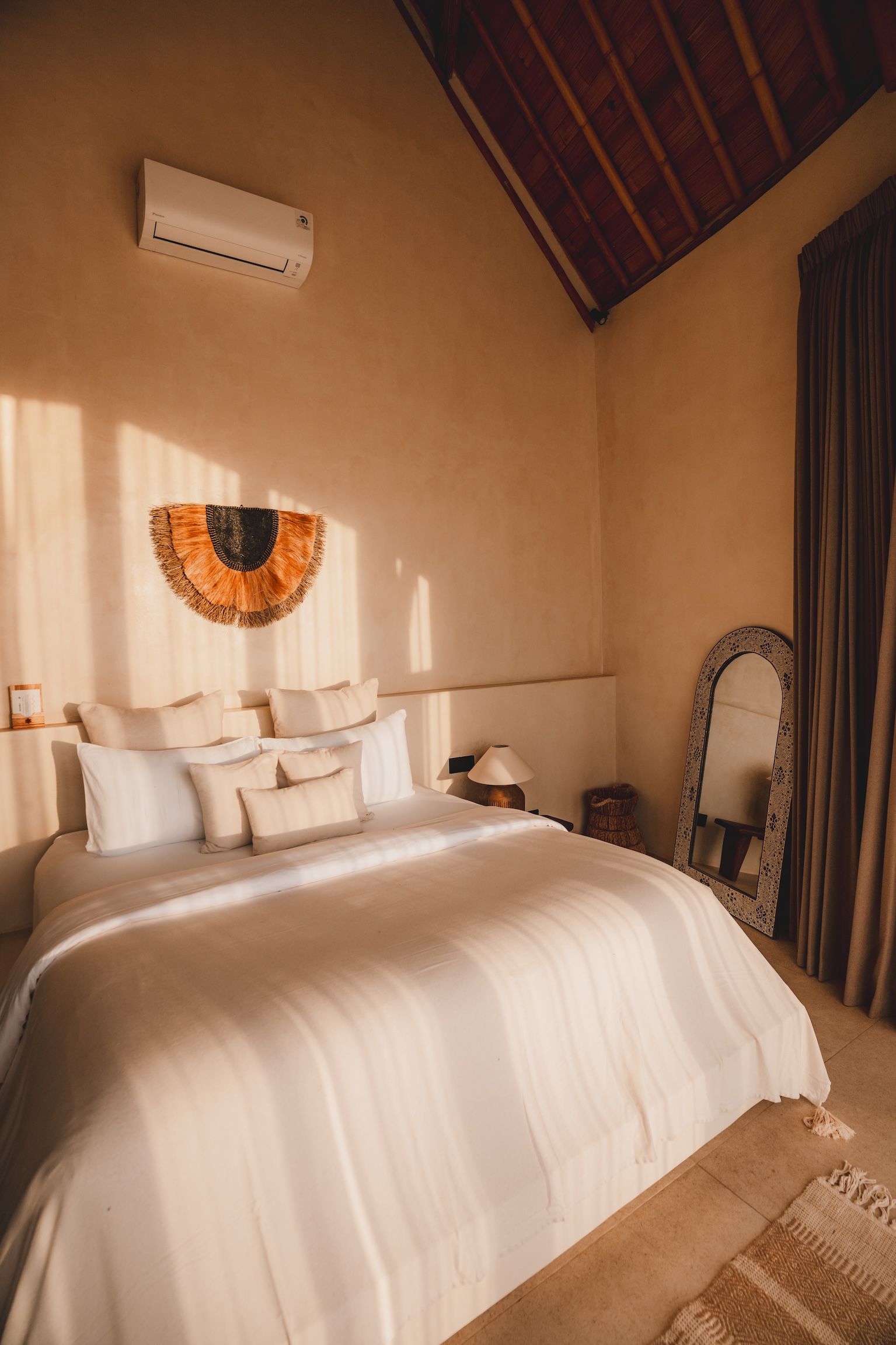 Cozy bedroom with a white bed, warm beige walls, woven decor, and sunlight streaming in from the left