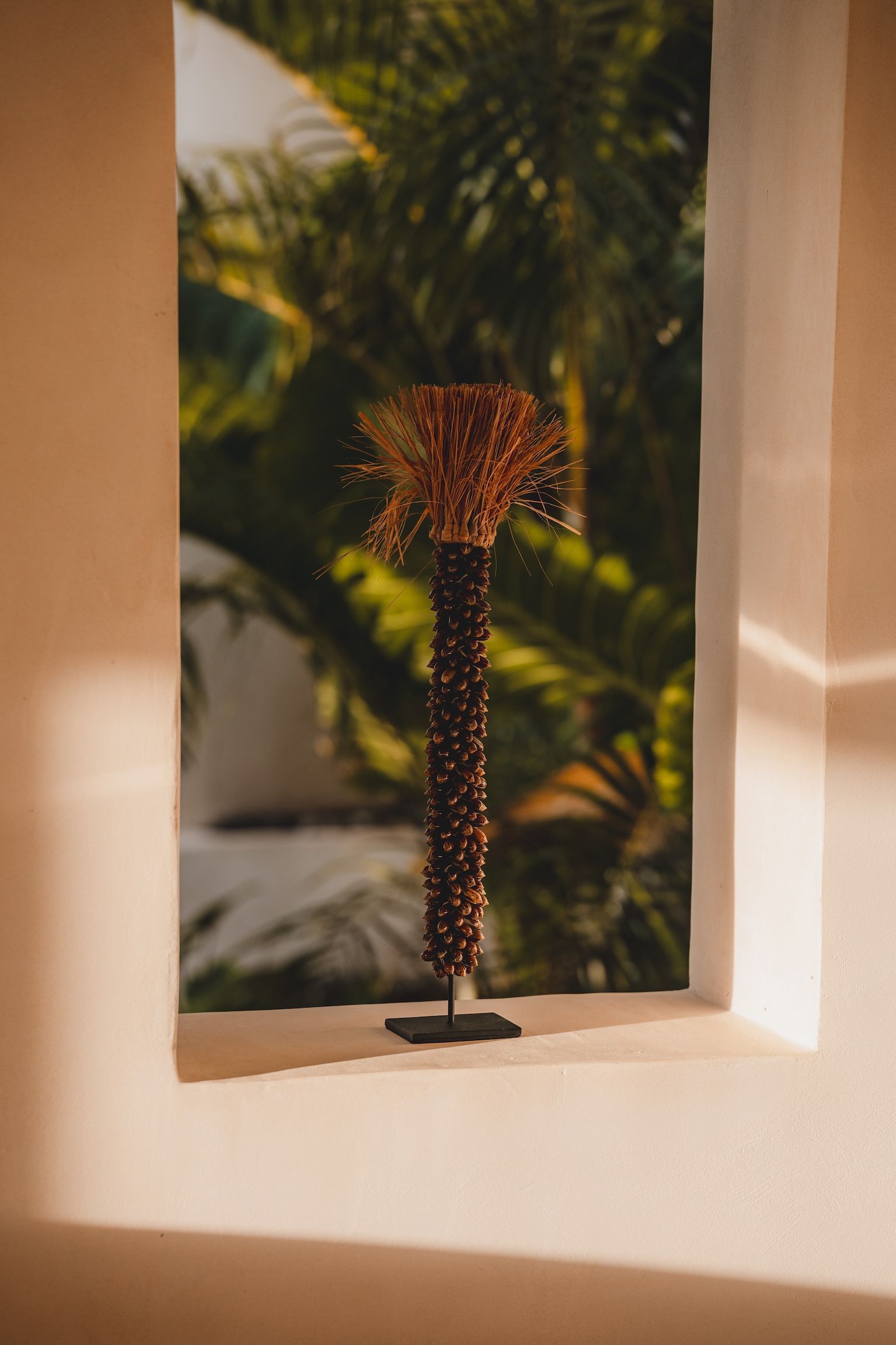 Decorative beaded sculpture on a windowsill, framed by a sunlit window with blurred palm leaves outside