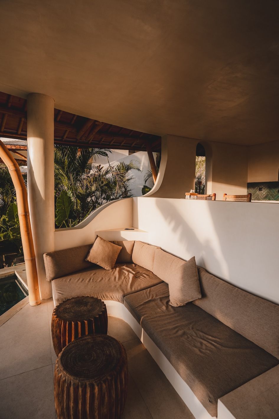 Sunlit outdoor lounge with tan sectional sofa, round tables, and white walls under a curved roof