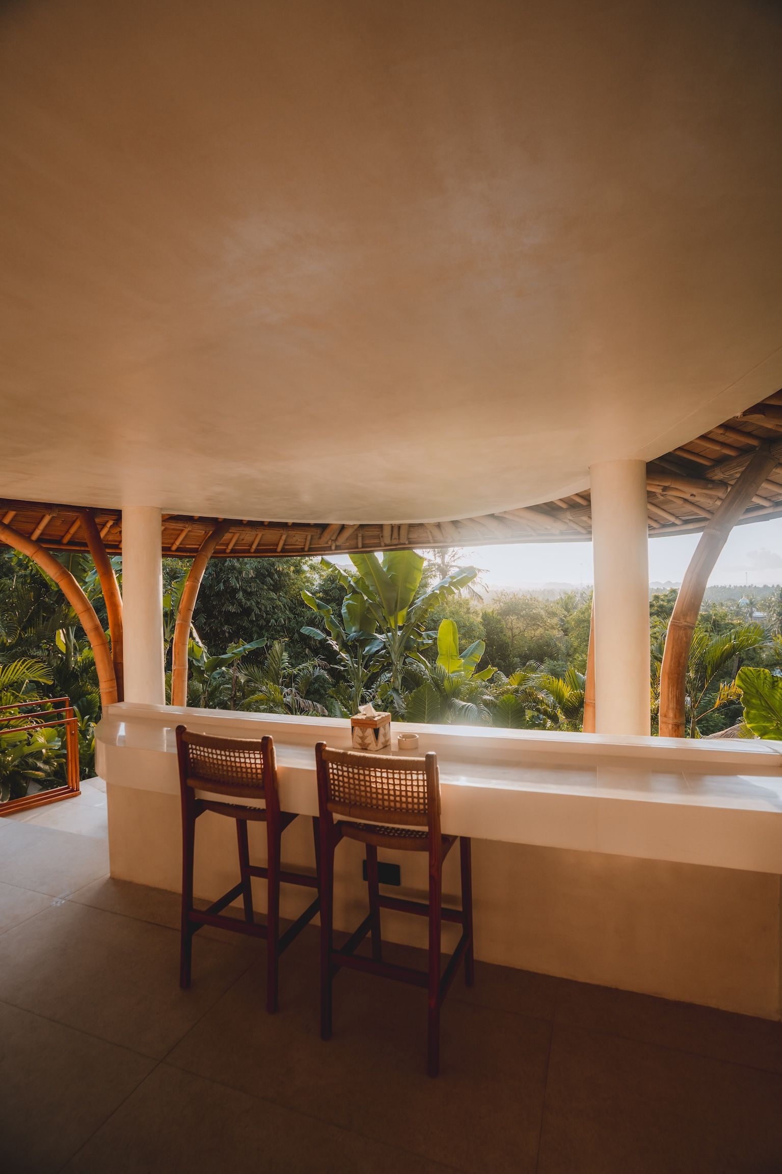 Tropical bar with two wooden stools overlooking lush greenery and sunlight beyond a shaded patio.