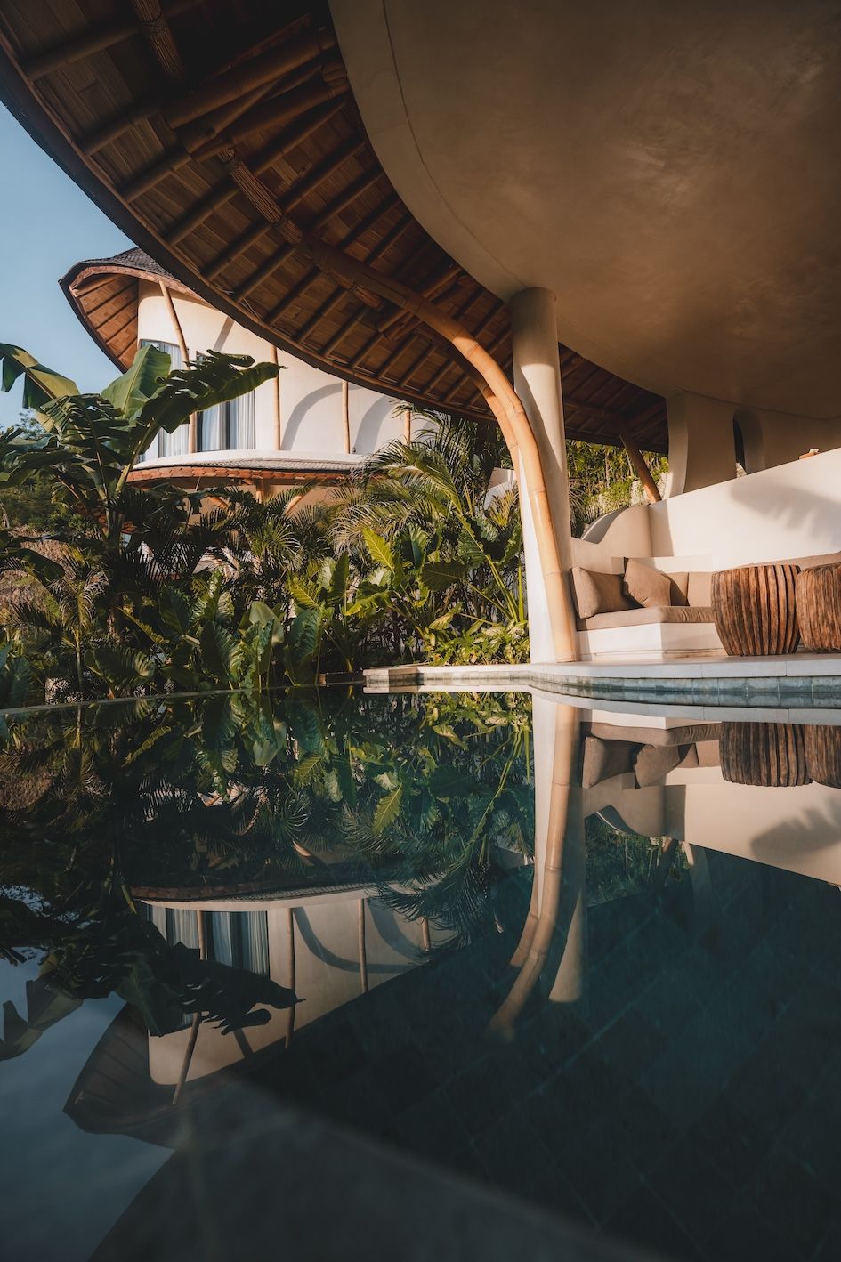 Tropical poolside resort with curved thatched roof, lush greenery, and calm reflective water at sunset