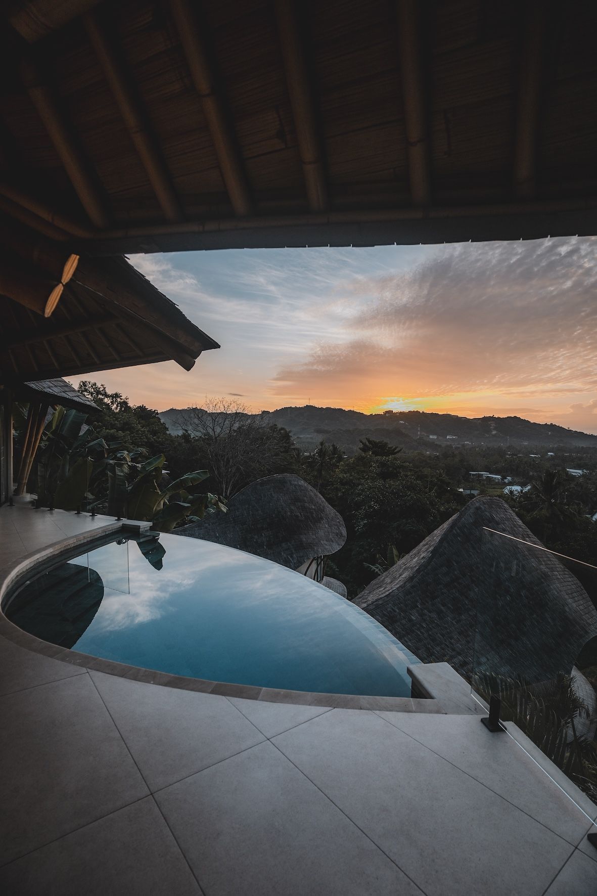 Infinity pool at sunset overlooking hills and thatched-roof huts from a shaded patio