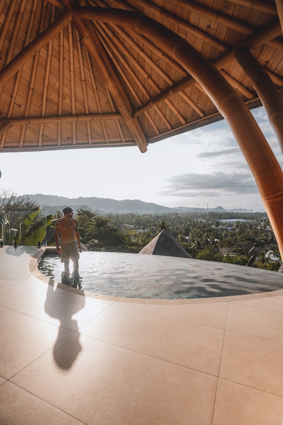 Two people stand by an infinity pool under a thatched roof, overlooking a sunlit valley.