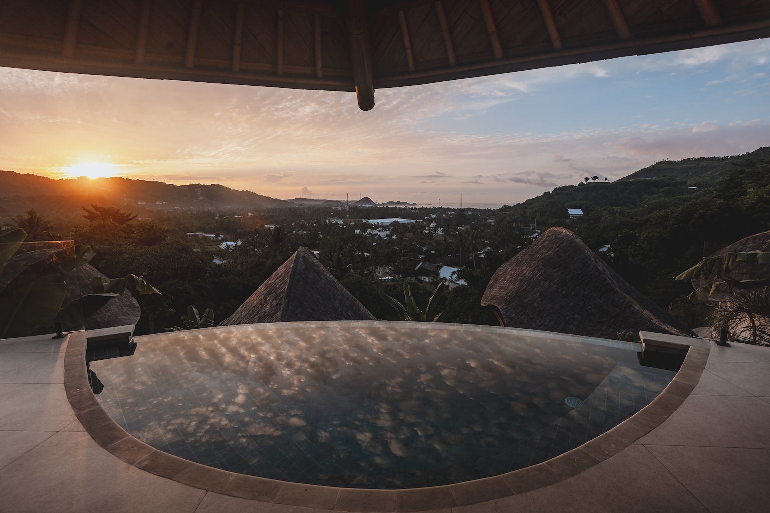 Sunset view from a thatched terrace over a valley, with a round infinity pool in the foreground