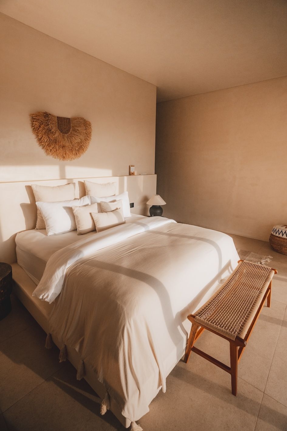 Neatly made bed in a beige bedroom with white linens, woven headboard decor, and a bench at the foot