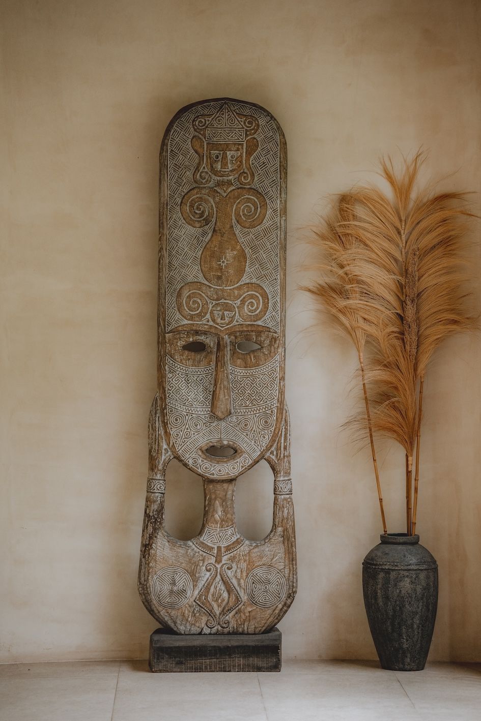 Tall carved wooden mask on a stand beside a vase of dried pampas grass against a beige wall