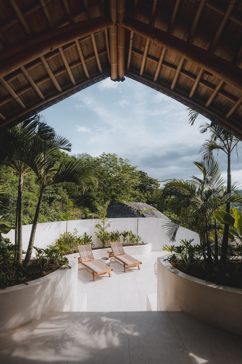 Thatched-roof patio overlooking lush tropical greenery and a valley, with wooden lounge chairs in the sun