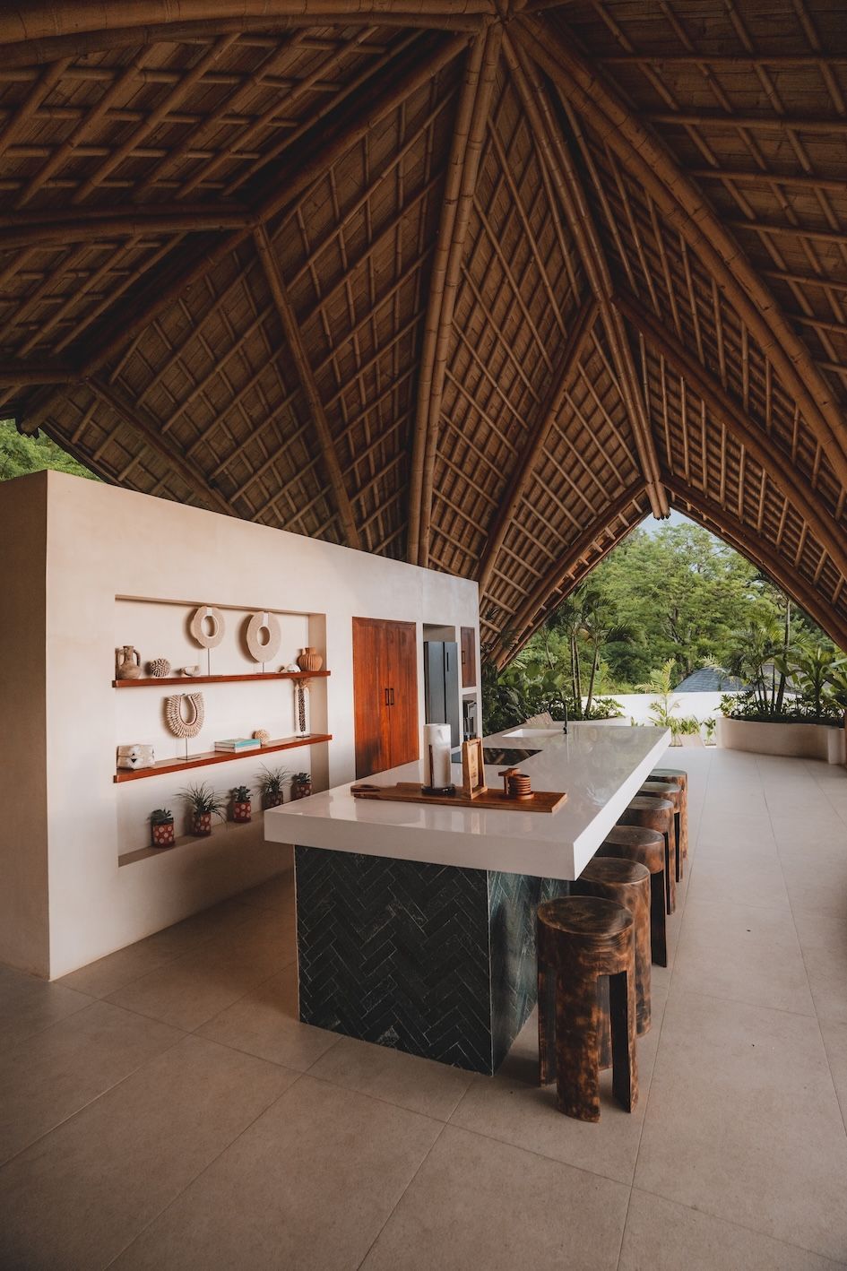 Open-air kitchen with a thatched roof, long island counter, and wooden shelves in a tropical setting
