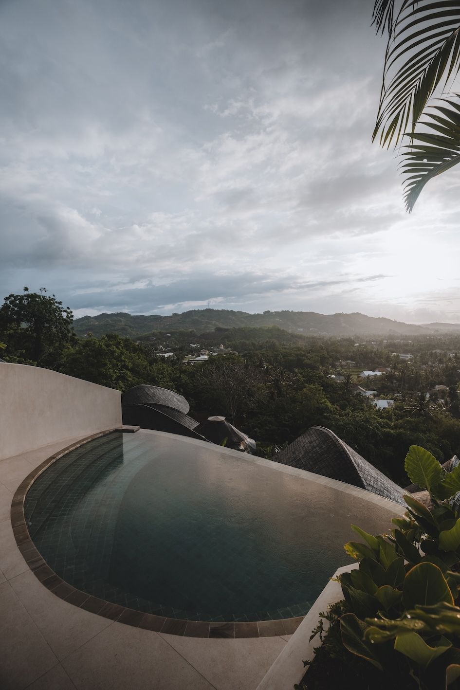 Infinity pool overlooking a lush valley at sunset, with cloudy sky and palm fronds framing the view