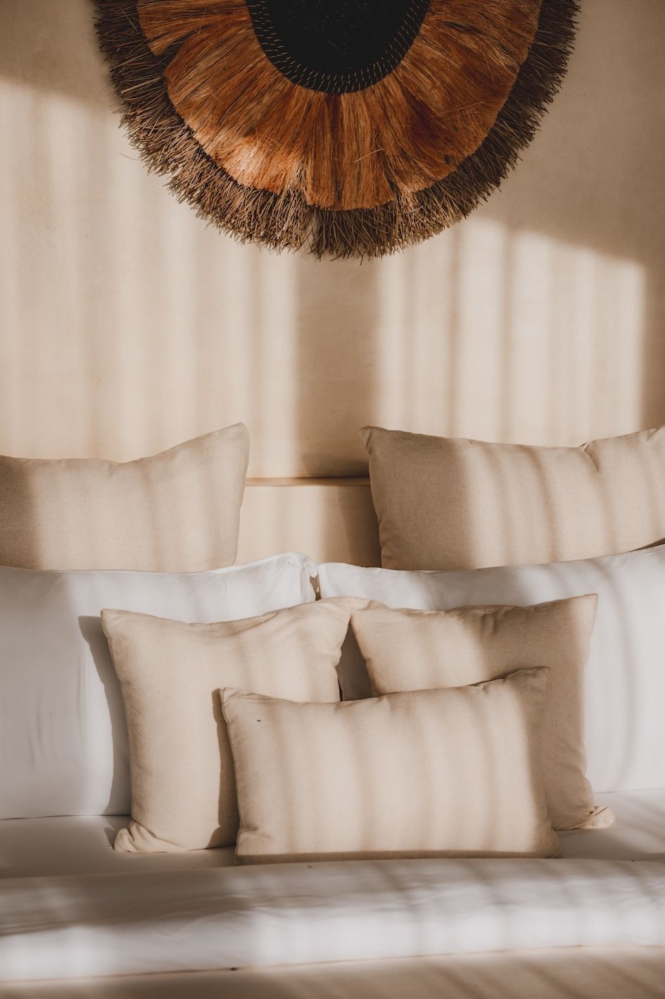 Sunlit beige bed with layered pillows beneath a large woven wall hanging