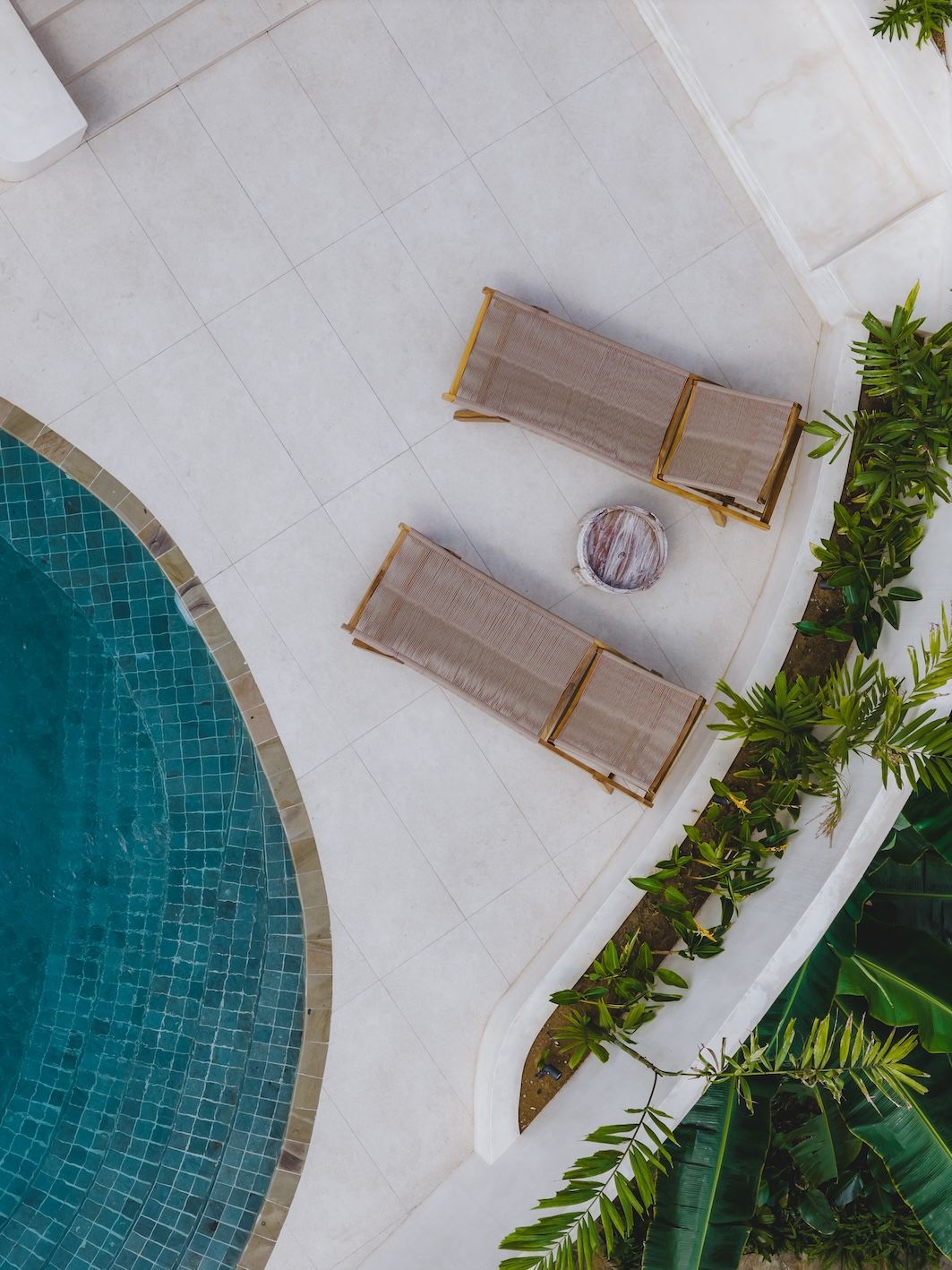 Overhead view of two lounge chairs beside a turquoise pool on a white patio with tropical plants