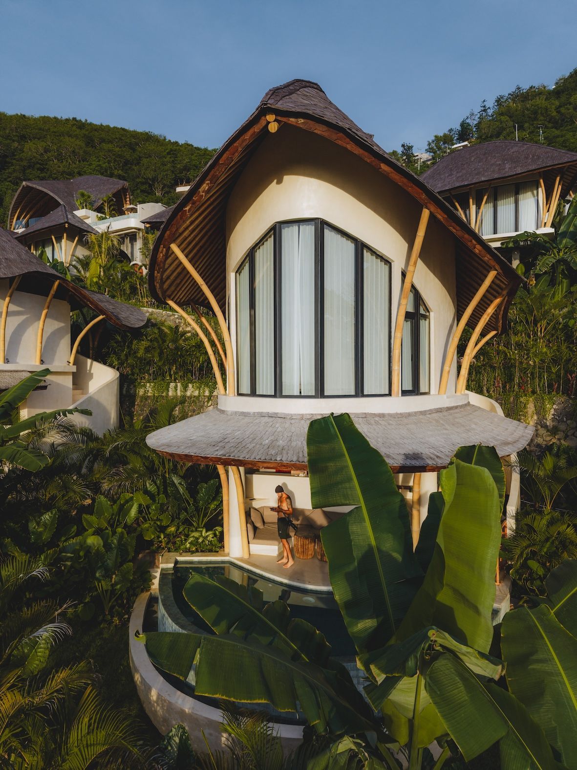 Modern tropical villa with curved roof, large windows, and lush greenery at dusk