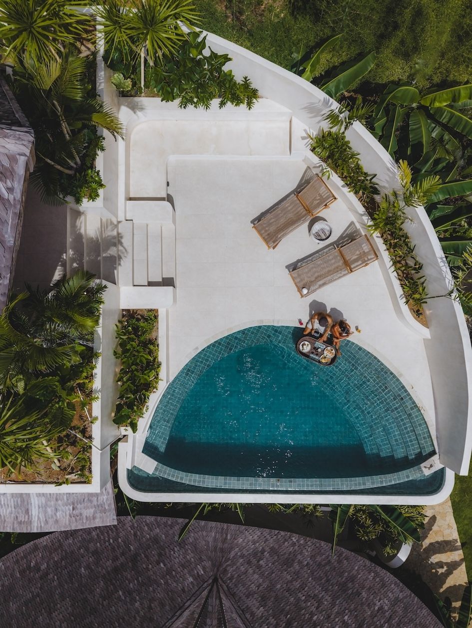 Overhead view of a white pool deck with a turquoise curved pool, lounge chairs, and tropical greenery.