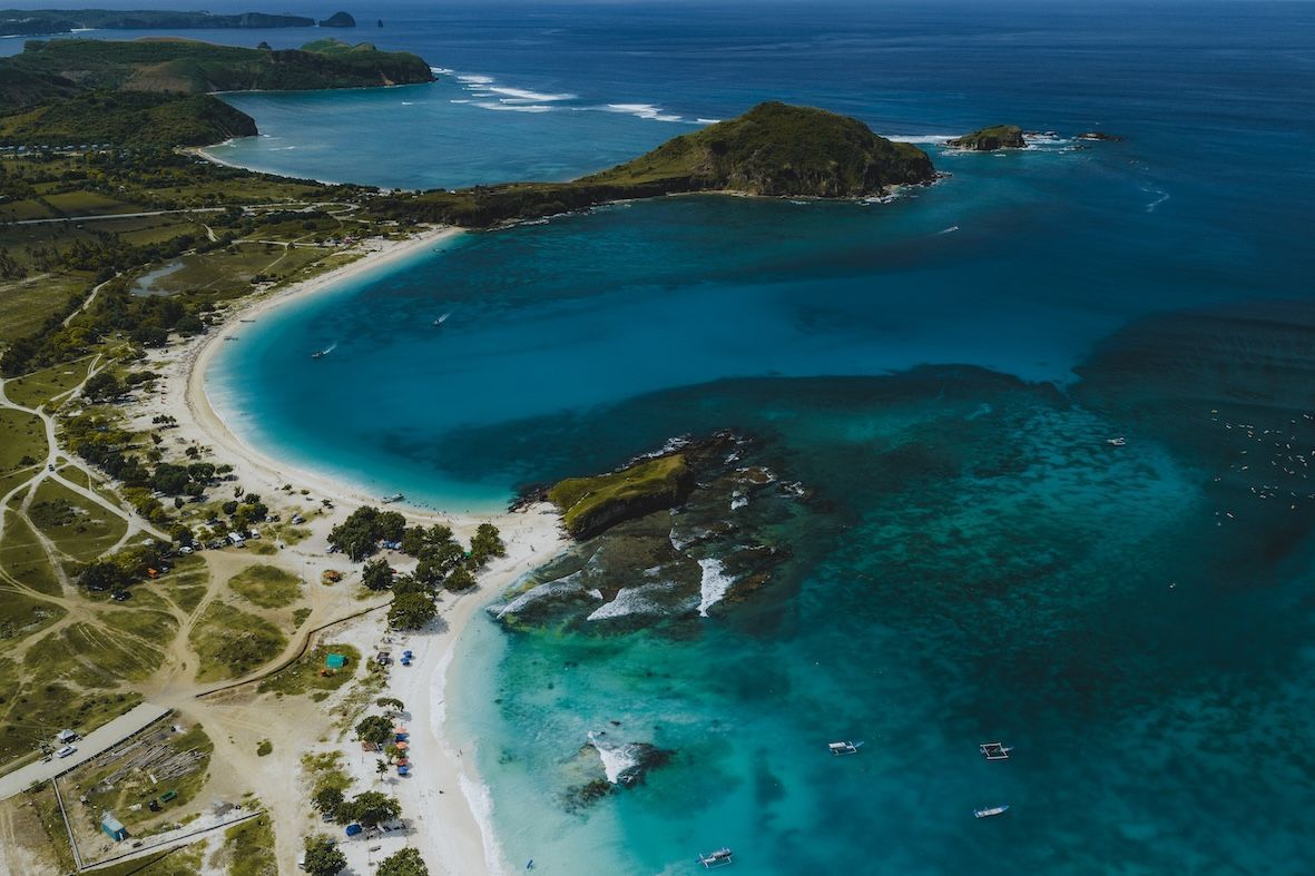 Aerial view of a turquoise bay with white sandy beach, rocky shoreline, and green coastal hills.