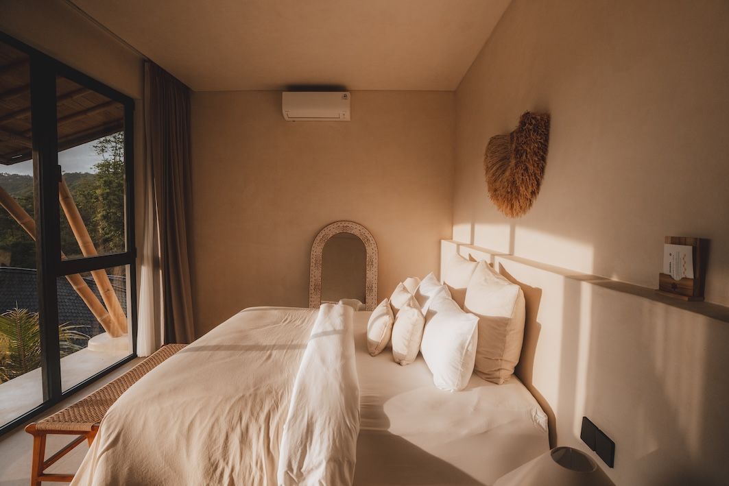Sunlit bedroom with a neatly made bed, wicker mirror, and large window overlooking greenery