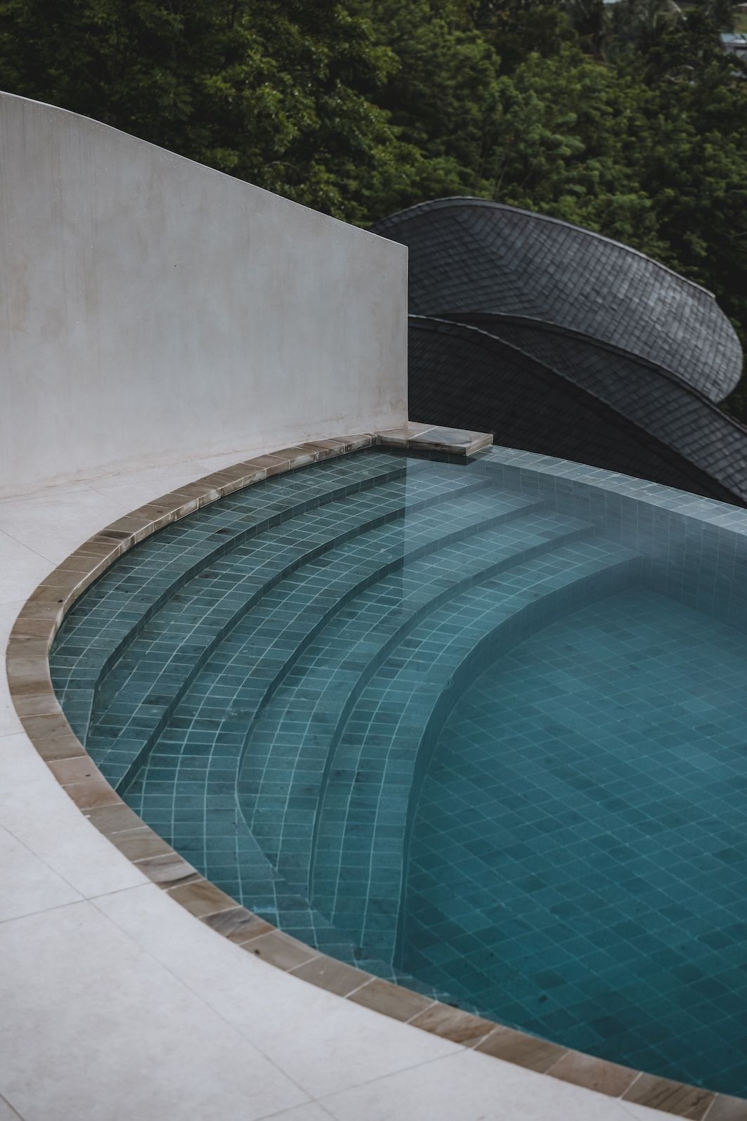 Curved blue pool with stepped ledge beside a white wall and dark woven lounger