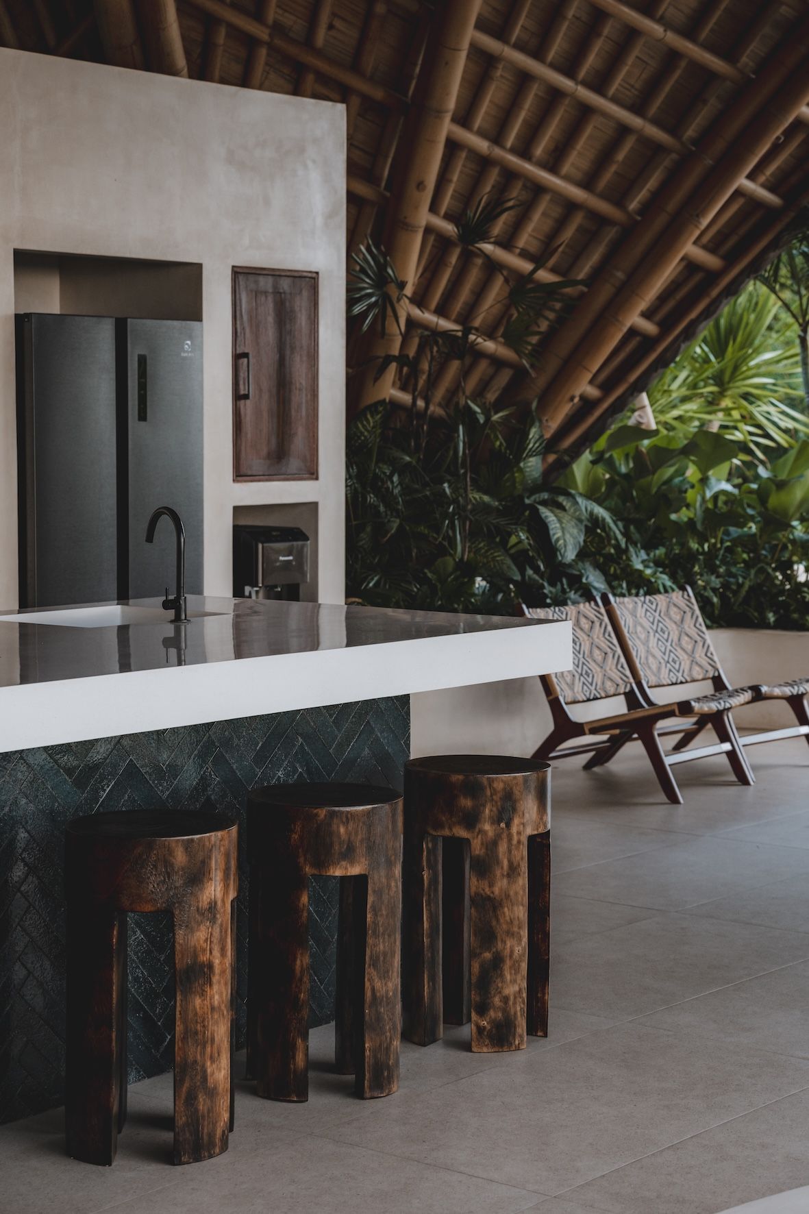 Modern tropical kitchen with white counter, dark wooden stools, and a thatched roof terrace view