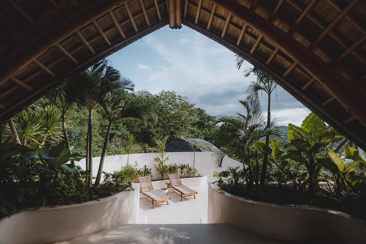 Tropical patio with wooden roof framing a white terrace, lounge chairs, and lush green trees beyond.