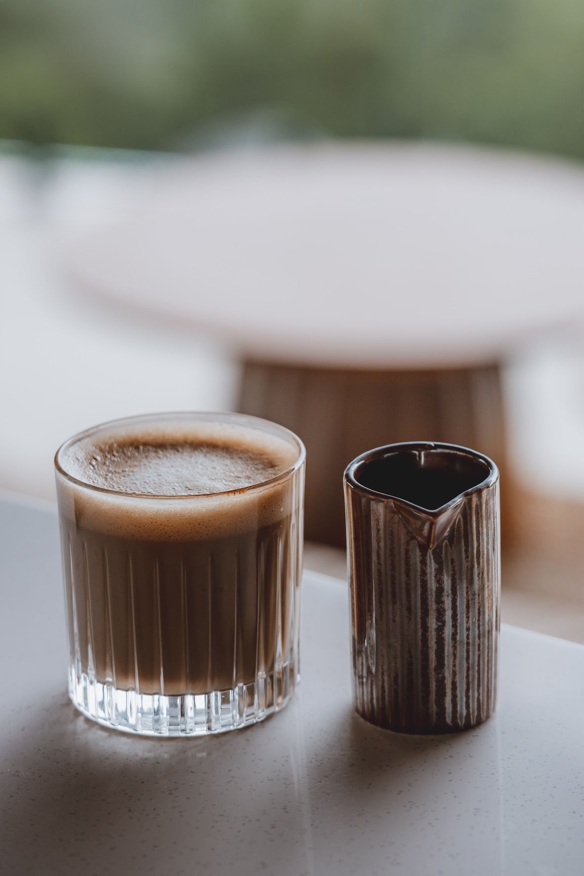 Two coffee drinks on a table, one in a glass and one in a dark metal cup, with a blurred chair behind them