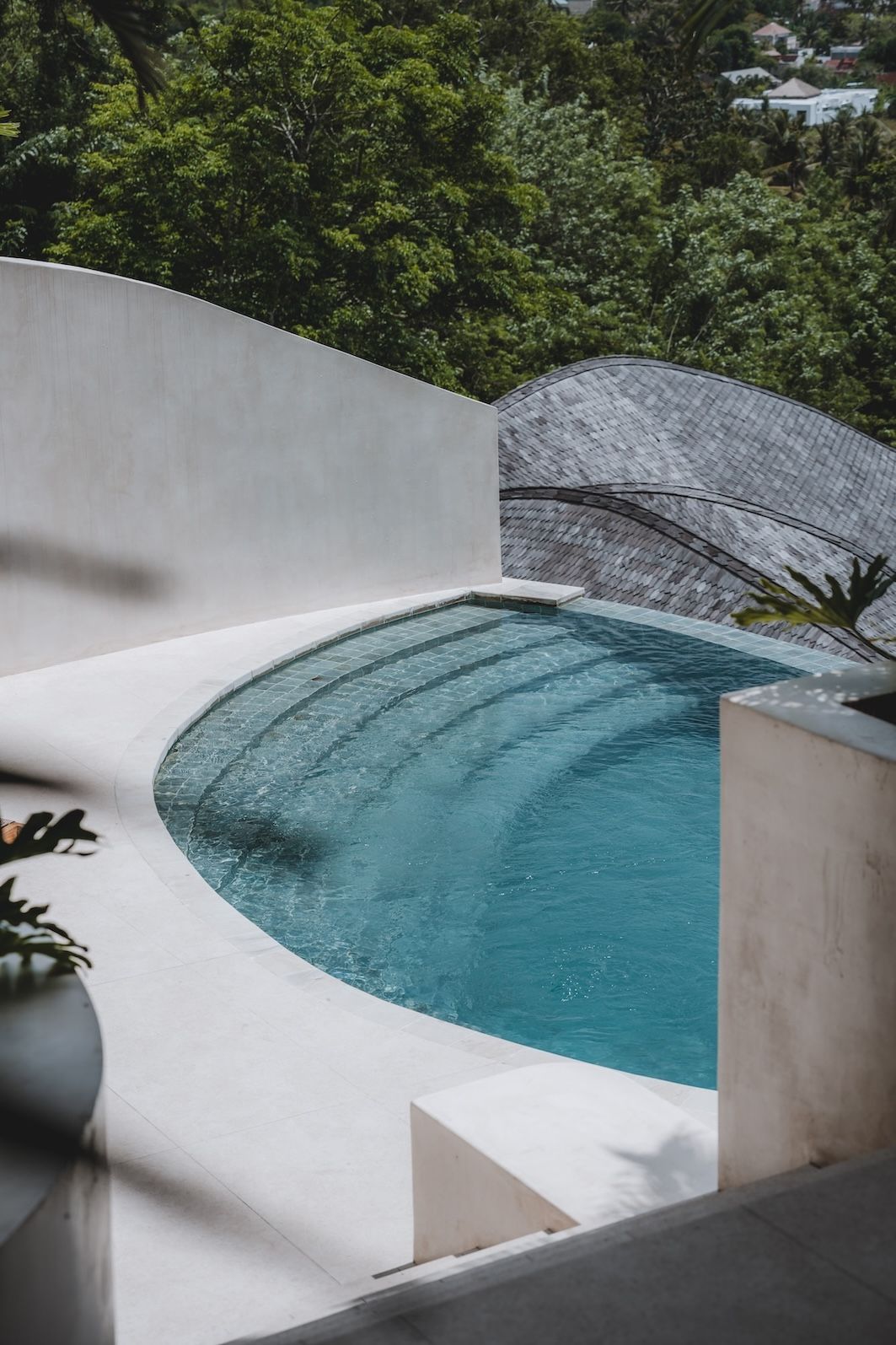 Curved turquoise pool beside a white patio wall, surrounded by lush green trees and a gray roofline.