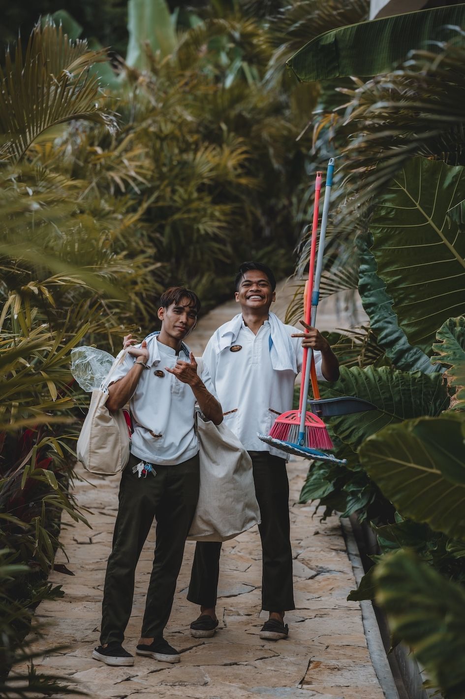 Two people walking on a sandy path through dense palm trees, one carrying a colorful pole.