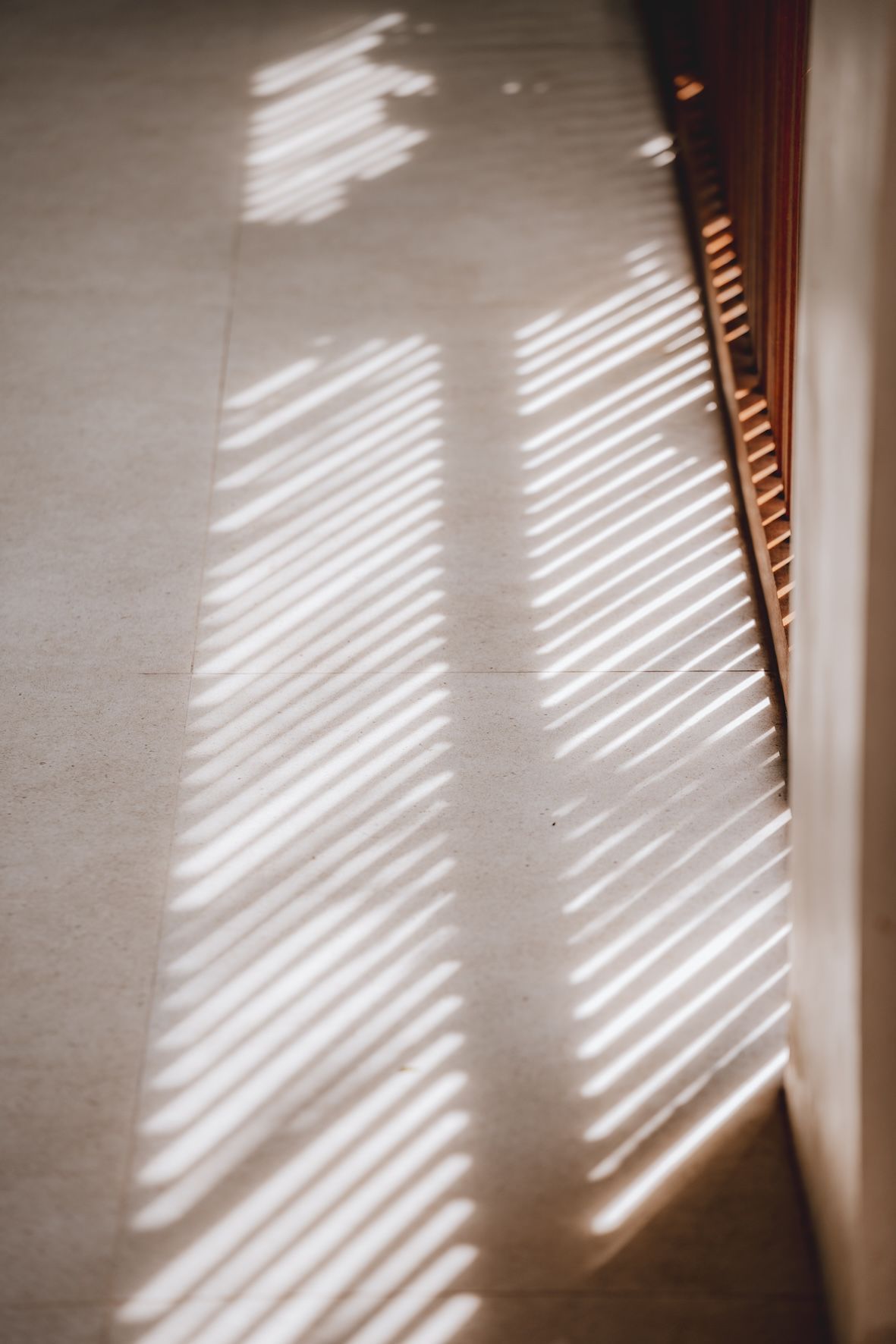 Sunlight through blinds casts diagonal shadows on a beige wall beside a window.