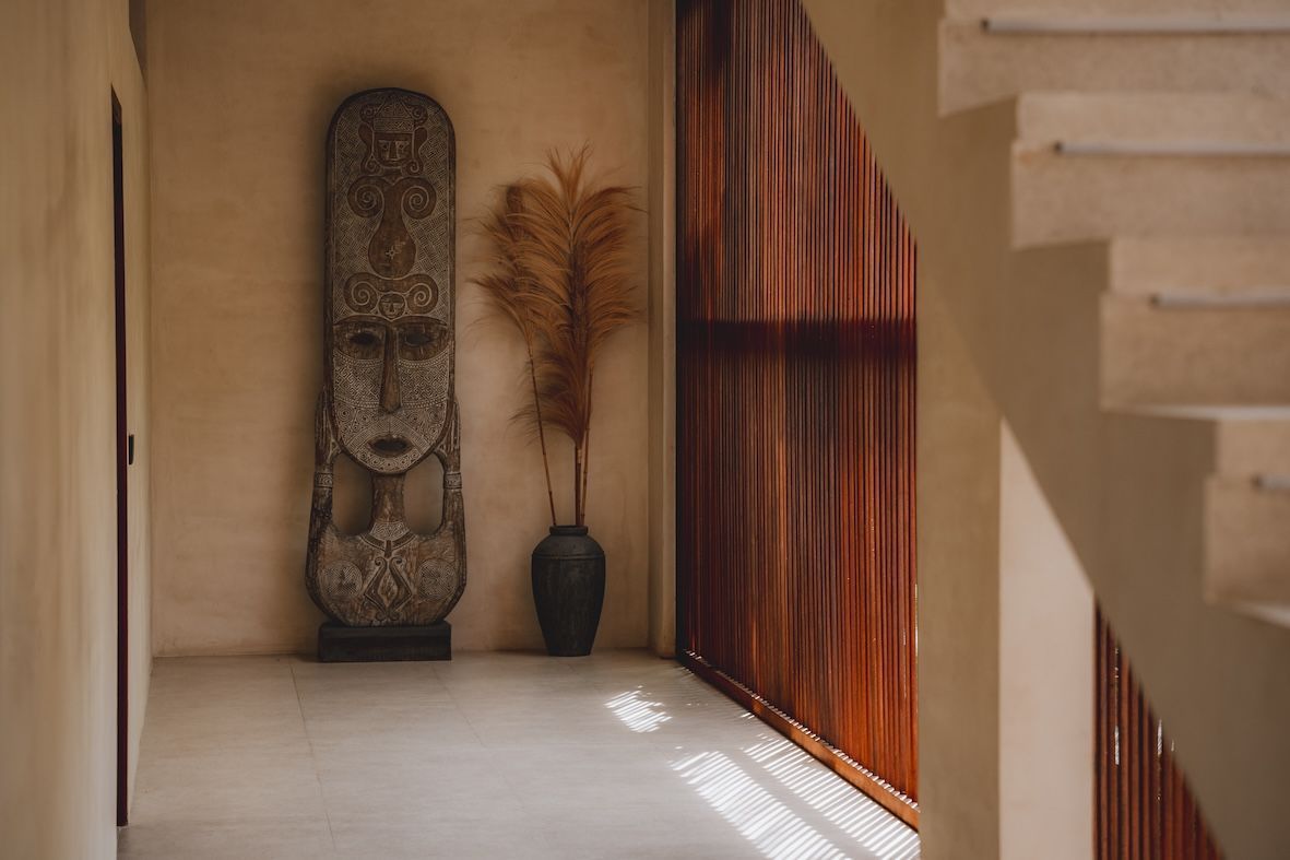 Minimalist hallway with a tall carved statue, vase, and warm light beside a wooden screen and staircase