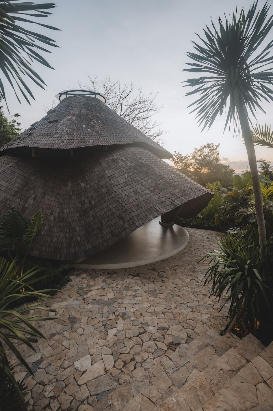 Thatched circular hut in a tropical garden at dusk, with stone path and tall palm-like trees.