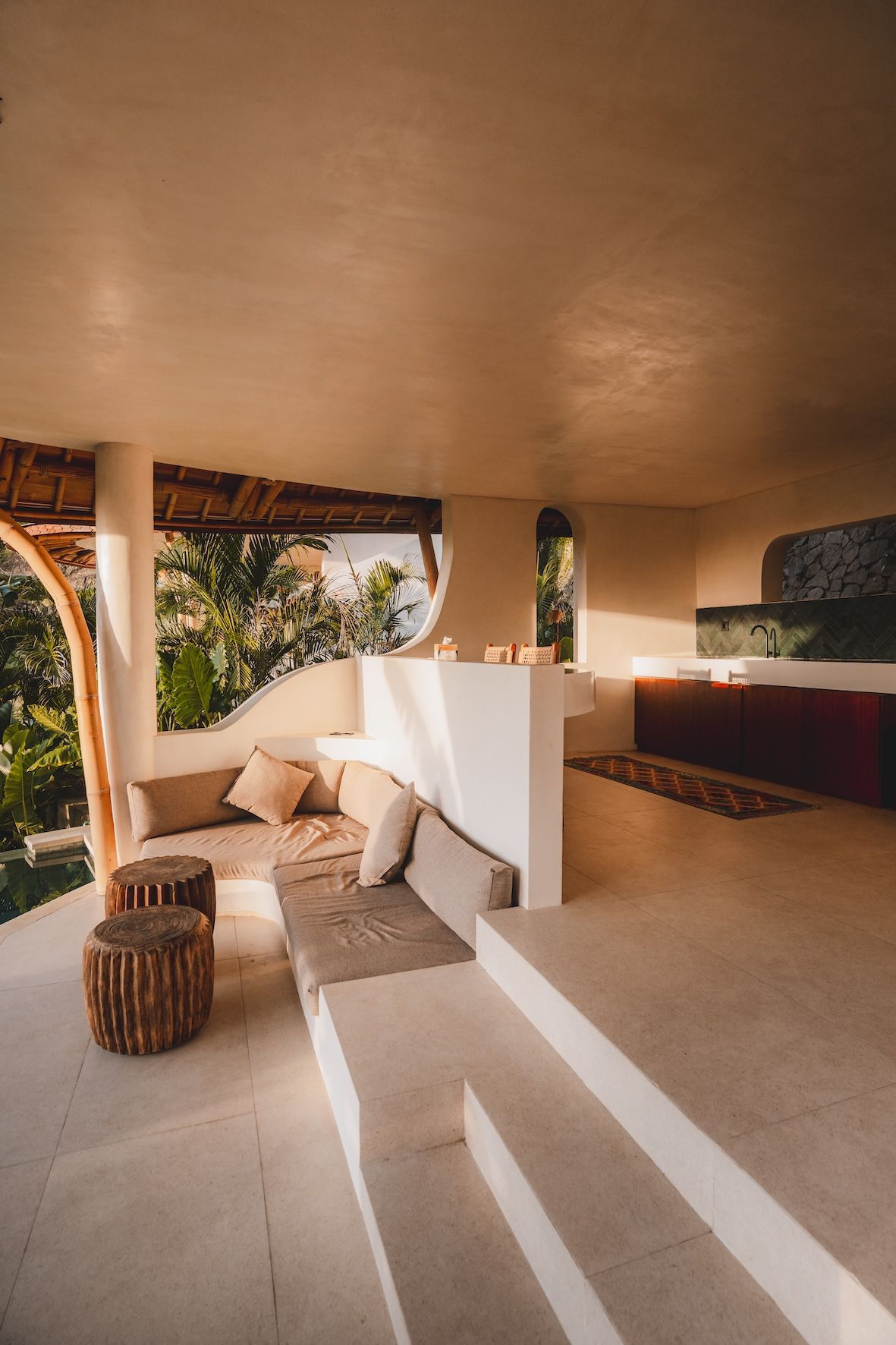 Sunlit beige lounge with wicker stools, cushions, and open-air view of greenery
